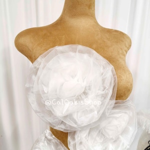 May include: A white sheer organza garment featuring large, layered, circular floral-shaped fabric rosettes. The piece is displayed on a tan mannequin and includes the text @CalOasisShop printed in white across the center of the fabric.