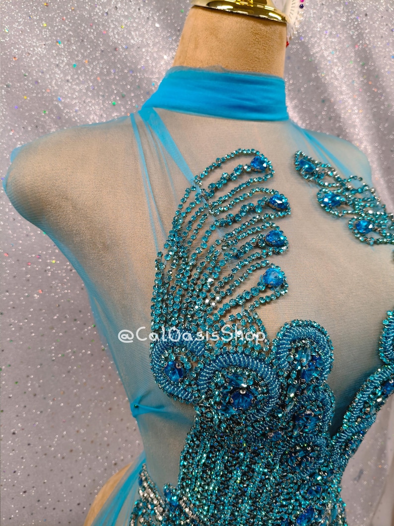 May include: A turquoise, sheer costume with intricate beadwork. The bodice features elaborate, beaded designs in shades of turquoise and blue. The costume has a high neckline and is displayed on a mannequin. The text "@ColOasisShop" is visible.