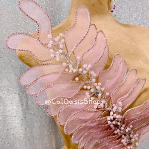 May include: A delicate, light pink decorative applique with a leaf-like design. The applique features a silver wire frame, embellished with small white beads and a touch of blue. The edges are trimmed with a shimmering pink material. The text "@CalOasisShop" is visible.