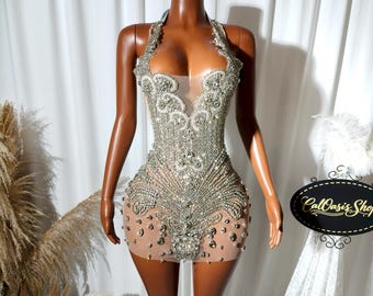 May include: A silver beaded halter dress with a sheer nude base. The dress features intricate beadwork and a plunging neckline. The dress is displayed on a mannequin. The CalOasis Shop logo is visible.