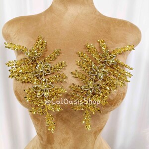 May include: Two golden, leaf-like appliques with crystal accents are displayed on a mannequin. The appliques are symmetrical, with a central floral design and radiating leaf patterns. The text "@CaLOasisShop" is visible on the mannequin.