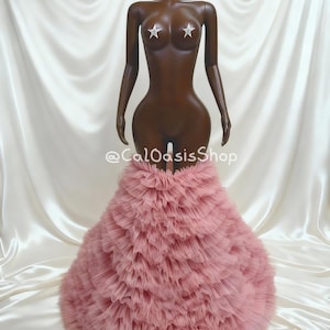 Puede incluir: Maniquí marrón con pezoneras en forma de estrella, luciendo una falda de tul rosa en capas. La falda tiene un diseño con volantes y se ensancha desde la cintura. El maniquí se encuentra sobre un fondo de satén blanco. El nombre de la tienda, "@CalOasisShop", es visible.