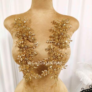 May include: Gold-toned floral applique with clear crystal accents. The applique is displayed on a mannequin. The text "@CaLOasisShop" is visible on the mannequin. The applique is likely for embellishing garments.