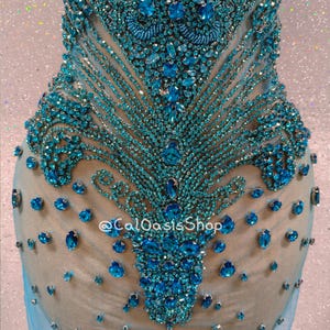 May include: A teal-colored, embellished garment with intricate beadwork and sparkling accents. The design features a central floral-like pattern with radiating lines of small beads and larger, jewel-toned stones. The garment is set against a neutral backdrop.