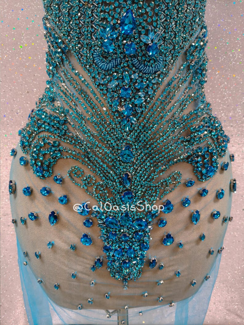 May include: A teal-colored, embellished garment with intricate beadwork and sparkling accents. The design features a central floral-like pattern with radiating lines of small beads and larger, jewel-toned stones. The garment is set against a neutral backdrop.