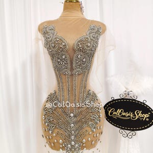 May include: A beige, sheer dress form adorned with elaborate silver beading and crystal detailing. The bodice showcases an intricate design, and the dress is displayed on a mannequin. The shop's logo, "CalOasis Shop," is visible.