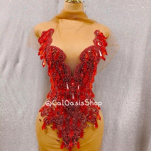 Könnte beinhalten: Ein rotes, mit Perlen besetztes Oberteil mit tiefem V-Ausschnitt. Das Oberteil ist mit roten Kristallen und Perlen in verschiedenen Größen verziert, was einen funkelnden Effekt erzeugt. Der Text "@CalOasisShop" ist auf dem Kleidungsstück zu sehen. Das Oberteil wird auf einer Schaufensterpuppe präsentiert.