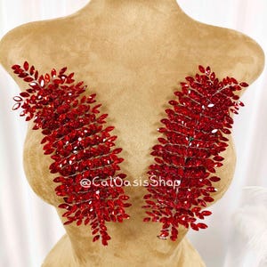 Puede incluir: Dos apliques simétricos en forma de hoja de cristal rojo. Cada aplique está compuesto por numerosos cristales rojos pequeños y facetados, dispuestos para imitar la forma de una hoja. Los apliques se muestran sobre un maniquí bronceado. El texto "@CalOasisShop" es visible.