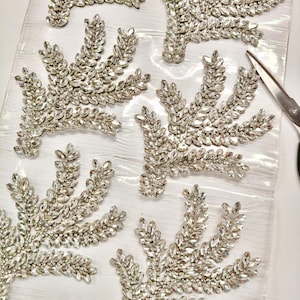 Puede incluir: Primer plano de varias aplicaciones de pedrería plateadas en forma de hoja. Cada aplicación está compuesta por numerosas piedras de estrás claras y brillantes, dispuestas para imitar un diseño de hoja estilizado. Un par de tijeras es visible en el fondo.