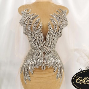 May include: A nude-colored bodice applique featuring an intricate arrangement of silver-toned crystals and beads in a leaf-like pattern. The design is symmetrical, covering the front of the garment with a deep V-neck shape and the text @CalOasisShop.