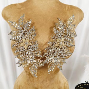 Puede incluir: Un par de apliques simétricos en tono dorado con cristales transparentes de corte marquesa dispuestos en un patrón de ramas con hojas. Cada pieza incluye un motivo central de flor de cinco pétalos con pequeños brillantes.