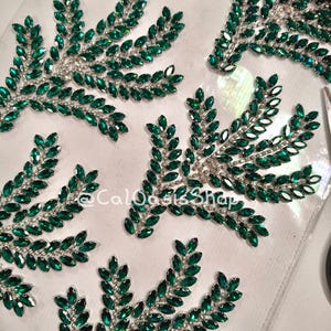 May include: Close-up of several emerald green rhinestone leaf appliques. Each leaf design features a silver-toned stem and is composed of numerous marquise-cut rhinestones. The appliques are arranged on a clear surface.