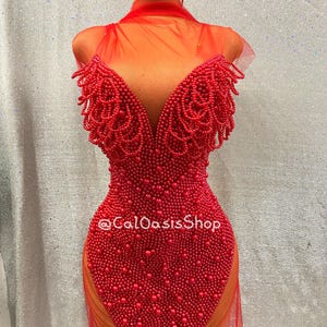 May include: A red beaded dress with a deep V-neck and sheer fabric. The dress is adorned with small, round beads and has a fitted shape. The text "@CalOasisShop" is visible on the dress.
