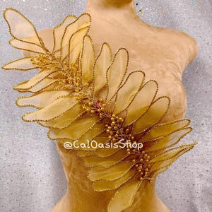 Peut inclure: Une applique en tissu de couleur dorée avec un motif de feuille et des accents perlés. L'applique est sur le torse d'un mannequin. Le texte "@CalOasisShop" est visible dans le coin inférieur droit de l'image.