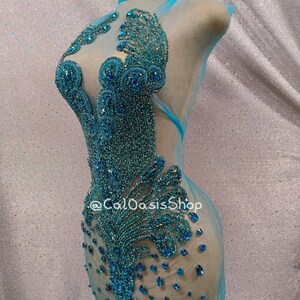 May include: A turquoise, sheer, embellished dress on a mannequin. The dress features intricate beadwork in shades of turquoise and blue, creating a detailed pattern. The dress has a high neck and a flowing, transparent skirt. The text "@CalOasisShop" is visible.