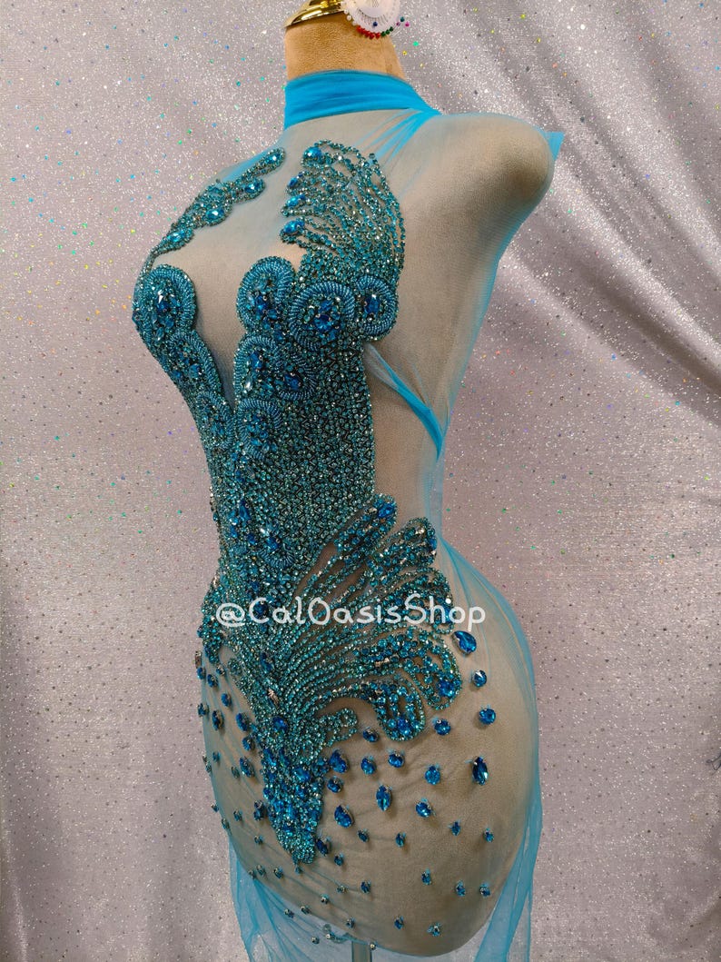 May include: A turquoise, sheer, embellished dress on a mannequin. The dress features intricate beadwork in shades of turquoise and blue, creating a detailed pattern. The dress has a high neck and a flowing, transparent skirt. The text "@CalOasisShop" is visible.