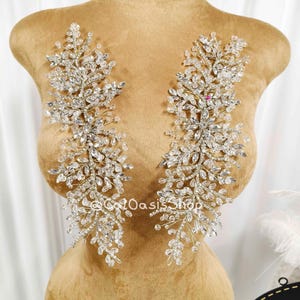 Może przedstawiać: Dwie srebrzyste aplikacje wysadzane kryształami, każda z kwiatowym i liściastym wzorem. Aplikacje są prezentowane na manekinie. Na zdjęciu widoczny jest tekst "@CatOasisShop".