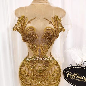 May include: A sheer, beige garment adorned with intricate gold beadwork in a floral design. The embellishments are concentrated on the bodice and extend downwards. The garment is displayed on a mannequin. The shop name "@CalOasisShop" is visible.
