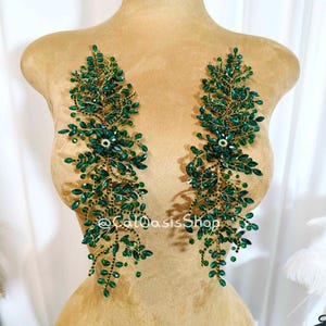 May include: Two emerald green beaded appliques in a leaf design. The appliques are attached to a mannequin. The leaves are made of green beads and gold wire. The appliques are symmetrical and are for clothing embellishment.