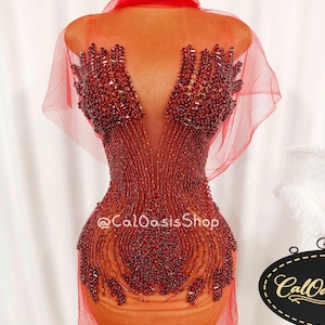 Puede incluir: Un vestido rojo con cuentas sobre un maniquí. El vestido presenta un escote en V profundo y patrones intrincados de cuentas. El vestido tiene una capa transparente y está diseñado para un evento formal. El texto "@CalOasisShop" es visible.