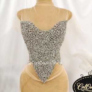 May include: A sheer beige bodysuit featuring a sweetheart neckline and a bodice densely covered in silver marquise-cut crystals. The garment has short sleeves and a pointed hemline. The text @CalOasisShop is visible on the crystal bodice.