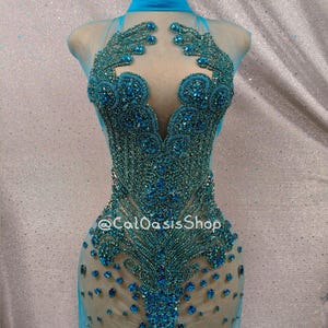 May include: A teal-colored, beaded dress on a mannequin. The dress features a high neckline and a sheer overlay. The bodice is heavily embellished with intricate beadwork, and the skirt is fitted. The text "@CalOasisShop" is visible on the dress.
