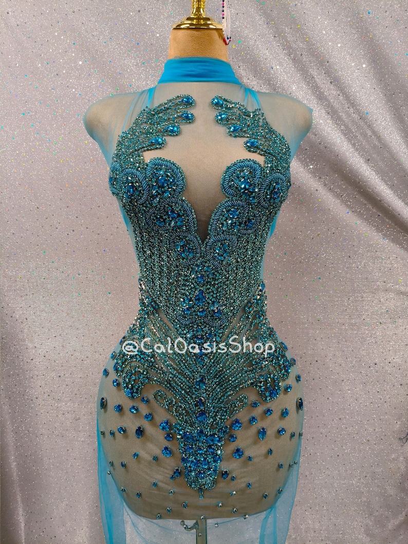 May include: A teal-colored, beaded dress on a mannequin. The dress features a high neckline and a sheer overlay. The bodice is heavily embellished with intricate beadwork, and the skirt is fitted. The text "@CalOasisShop" is visible on the dress.