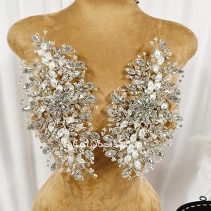 Puede incluir: Dos adornos florales plateados con detalles de cristal transparente. El diseño presenta detalles en forma de hojas y piedras brillantes, creando un aspecto glamuroso. Los adornos se exhiben en un maniquí.