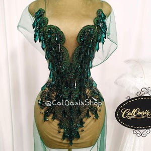 Może przedstawiać: Zielona, ​​ozdobiona sukienka z głębokim dekoltem w kształcie litery V i przezroczystą nakładką. Sukienka posiada skomplikowane koraliki i kryształowe akcenty w odcieniach zieleni. Sukienka jest prezentowana na manekinie.