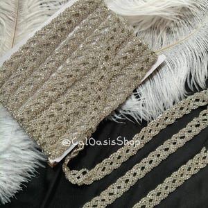 May include: A roll of silver rhinestone trim with a woven design. The trim is displayed on a white surface with white feathers and a black background. The text "@ColOasisShop" is visible.