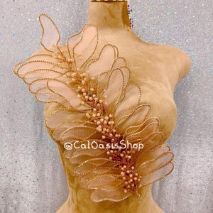 May include: A gold-toned applique with a leaf-like design. The applique features sheer, petal-shaped elements outlined with gold beading and adorned with small, pearl-like beads. The text "@CalOasisShop" is visible on the mannequin.