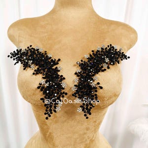 May include: Two black beaded appliques with silver rhinestone accents are displayed on a tan dress form. The appliques are symmetrical, with a floral-like design. The dress form has a gold-colored top and is set against a white background.