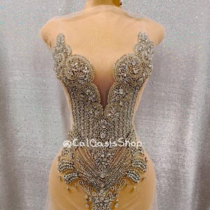 Puede incluir: Un corpiño beige con intrincados adornos de cristal plateado y transparente. El diseño presenta un escote en V profundo y un patrón detallado de piedras brillantes, con el texto "@ColOasisShop" visible.