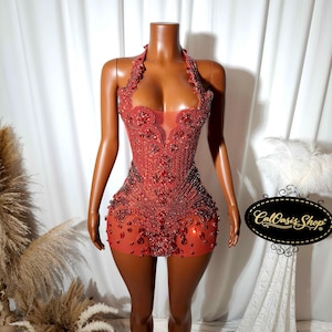 May include: A red, halter-neck mini dress adorned with intricate beadwork and embellishments. The dress features a sheer panel at the bust and is displayed on a mannequin. The dress is a vibrant shade of red.