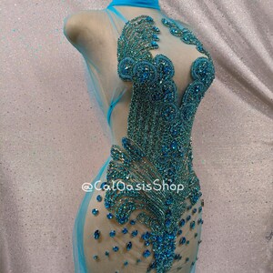 May include: A turquoise dance costume on a mannequin. The sheer fabric is adorned with intricate beadwork in shades of blue, creating a dazzling effect. The costume features a high neck and a flowing skirt. The text "@CalOasisShop" is visible.