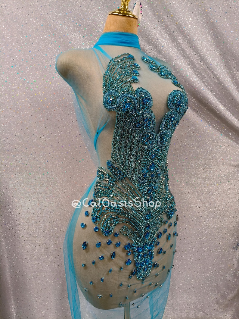 May include: A turquoise dance costume on a mannequin. The sheer fabric is adorned with intricate beadwork in shades of blue, creating a dazzling effect. The costume features a high neck and a flowing skirt. The text "@CalOasisShop" is visible.