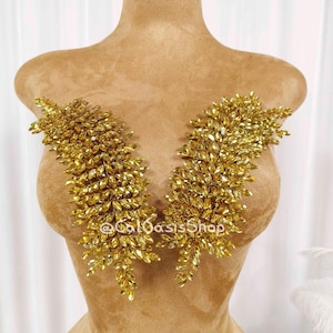 Puede incluir: Dos apliques dorados en forma de hoja, densamente cubiertos de pequeños cristales brillantes, se exhiben en un maniquí. Los apliques son simétricos y están colocados para adornar el busto. El maniquí es de color beige neutro.