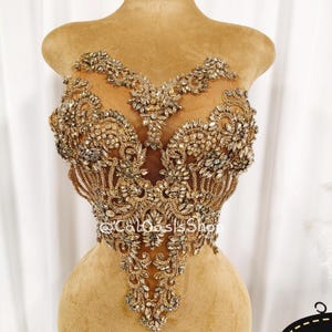 Może przedstawiać: Złoty gorset z misternymi koralikami i kryształowymi zdobieniami. Projekt charakteryzuje się centralnym wzorem z głębokim dekoltem i delikatnymi detalami. Gorset jest prezentowany na manekinie, prezentując swoje bogate rzemiosło.