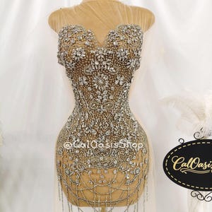 Pode incluir: Um vestido sem mangas em tule transparente cor de pele, com uma densa sobreposição de cristais e strass prateados em padrões florais e geométricos. O corpete tem um decote em coração e a bainha inclui franjas de cristal pendentes.