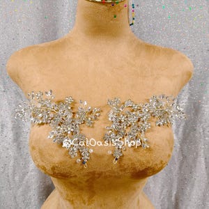 May include: A tan dress form displays two symmetrical, ornate crystal and silver appliques. The appliques are floral in design, with clear crystals and silver metal. The text "CalOasisShop" is visible on the dress form.