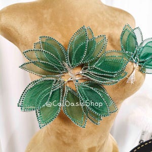 Può includere: Un accessorio floreale decorativo con petali di tessuto verde a strati, finiture argentate e piccoli abbellimenti scintillanti. Il design è disposto a forma di fiore ed è attaccato a un manichino. Il testo "@CalOasisShop" è visibile.