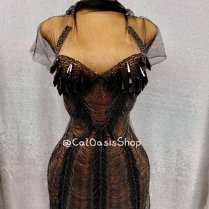 Peut inclure: Robe noire à perles sur mannequin. La robe présente un col licou, un corsage transparent et une silhouette ajustée. Le corsage est orné de perles sombres et d'éléments pendants. La jupe est faite de brins perlés. Le texte "@CalOasisShop" est visible.