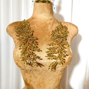 May include: Two gold-coloured, leaf-shaped appliques with sparkling embellishments are displayed on a mannequin. The appliques are symmetrical, with a central design and branches extending outwards. The mannequin is a warm tan colour, and the background is a white curtain.