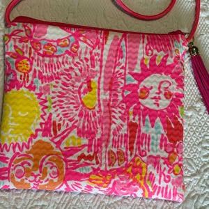May include: A vibrant, patterned crossbody bag with a pink, yellow, and orange floral design. The bag features a pink zipper, a pink tassel, and a pink cord strap. The bag is approximately 8 inches wide and 9 inches tall.