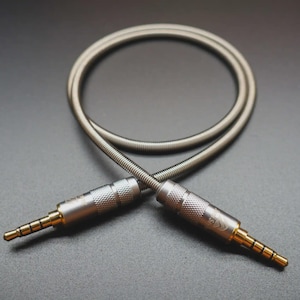 Puede incluir: Cable de audio plateado con dos conectores macho de 3,5 mm. El cable está enrollado y tiene un diseño trenzado. Los conectores tienen un agarre texturizado y puntas doradas. El fondo es gris oscuro.