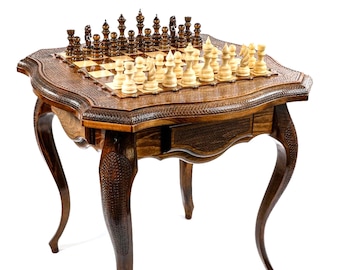 May include: A detailed, wooden chess set with a matching table. The chess pieces are arranged on a checkered board. The table has a drawer and ornate, curved legs. The wood has a rich, dark brown finish, and the table is set against a white background.