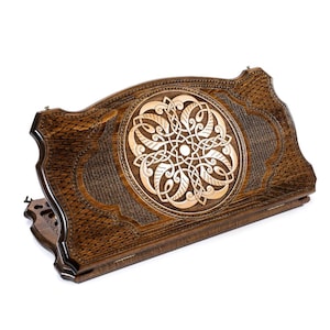 May include: A wooden backgammon board with intricate carved details. The board is primarily brown with a lighter wood inlay featuring a circular Celtic knot design. The edges have a textured pattern, and the board is slightly angled.