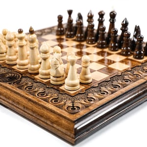 May include: A wooden chess set with intricately carved detailing around the board's perimeter. The chess pieces are arranged on a checkered board, with light and dark brown pieces set up for play. The set is displayed on a white background.