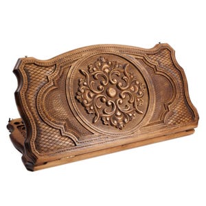 May include: A wooden backgammon board with intricate carvings. The board is a warm brown color and features a central floral design. The edges have a diamond pattern. The board is open, revealing the playing surface.