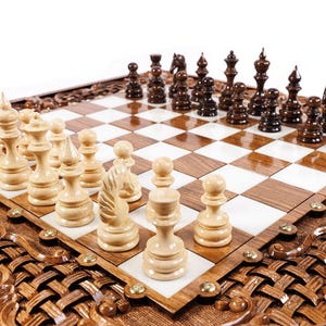 May include: A wooden chess set with intricately carved detailing around the board's perimeter. The chessboard features alternating light and dark squares, with light-colored and dark-colored chess pieces arranged for play. The set is displayed on a white background.